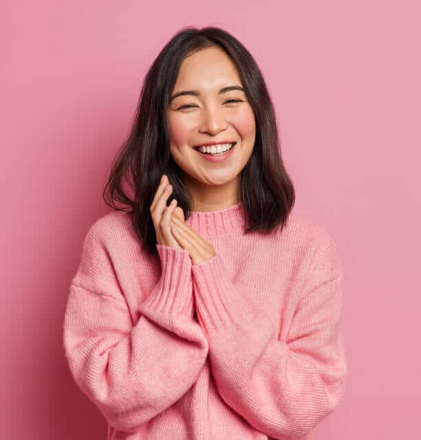 A young woman with medium-length dark hair smiles brightly, wearing a cozy pink sweater. She stands against a solid pink background, hands clasped near her face, looking cheerful and happy.