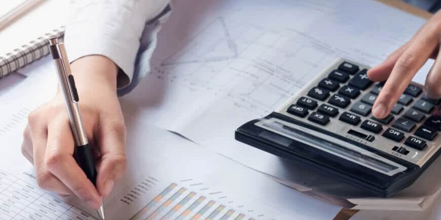 A person uses a calculator and writes on financial documents with charts and graphs on a desk, suggesting work related to accounting or data analysis.